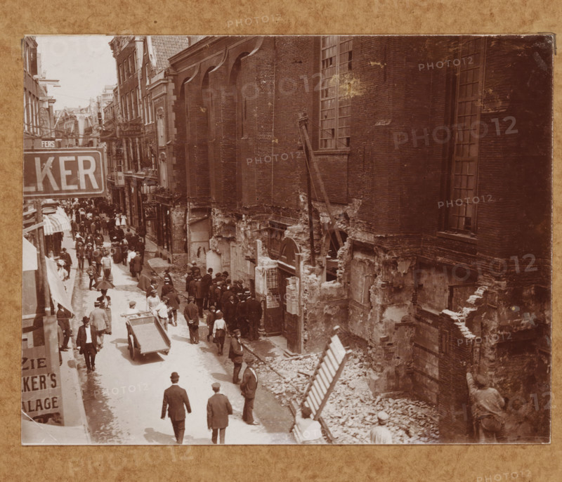 Street view with the Nieuwezijds Chapel in Amsterdam during the demolition in 1908 (former title), View of the Kalverstraat with the Nieuwezijds Chapel in Amsterdam during the demolition in 1908, photograph, annotation: ''08.150, 446', annotation: 'NZ Chapel', Amsterdam, 1908, photographer, paper, pasteboard, daylight collodion silver print, height 118 mm x width 154 mm, height 369 mm x width 295 mm, street (city(-scape) with figures, staffage), church (exterior) - QQ - small church, chapel, Kalverstraat, Nieuwezijds Chapel, A sepia street scene framed by tall brick buildings shows a busy thoroughfare with many pedestrians wearing period clothing and hats, some carrying umbrellas and others pushing a handcart. The left side features shopfronts and awnings lining the narrow street while the right side displays a large, weathered brick wall with tall windows and exposed, crumbled lower masonry revealing rubble and construction debris. A group of people congregates near a doorway set into...