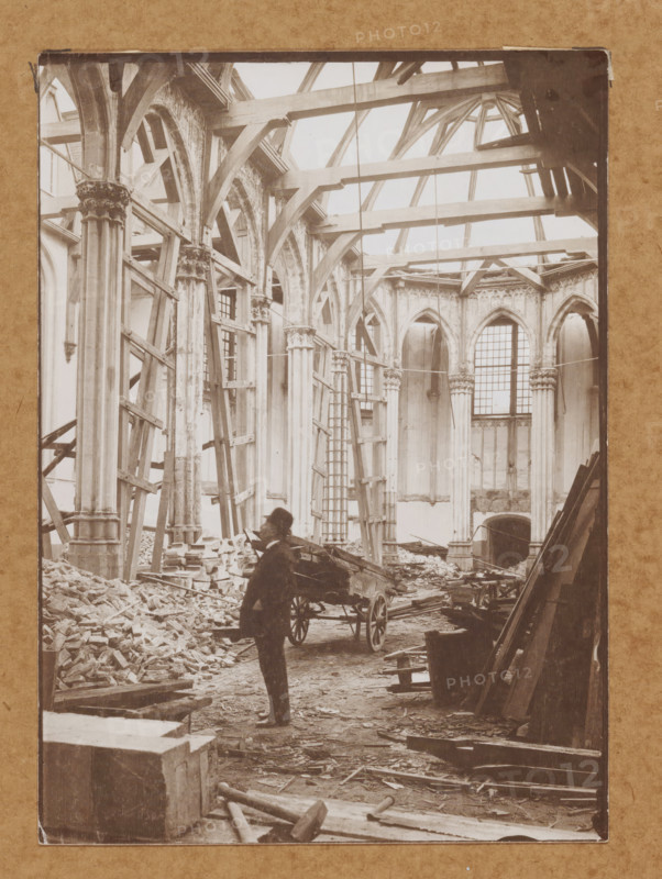 Interior of the Nieuwezijds Chapel in Amsterdam during the demolition in 1908, with Johannes van Tetterode, photograph, Amsterdam, 1908, photographer, paper, pasteboard, daylight collodion silver print, height 178 mm x width 128 mm, height 368 mm x width 293 mm, Person, Johannes van Tetterode, interior of church, Nieuwezijds Chapel, A large, partially ruined stone interior with tall, ornate columns and pointed arches, many wooden beams and scaffolding across the upper space, broken masonry and rubble piled on the floor, a wooden cart with large wheels near the center, a standing figure in dark clothing beside the cart, ladders leaning against columns, scattered planks and tools on the ground, and tall windows with segmented panes set into the far wall.