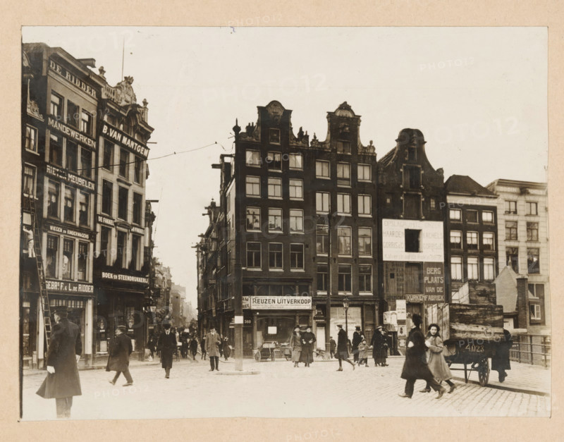 View of Sophiaplein or Muntplein in Amsterdam, photograph, annotation: ''17.2, Gift from photographer C. Steenbergh, Sophiaplein, On the right 5 houses being demolished', annotation: 'Sophiaplein (= Muntplein)', photographer: Cornelis Johannes Steenbergh (possibly), Amsterdam, 1900 - 1917, Cornelis Johannes Steenbergh, photographer, paper, pasteboard, gelatin silver print, developed gelatin silver print, height 126 mm x width 172 mm, height 183 mm x width 237 mm, square, circus, etc. (city(-scape) with figures, staffage), Muntplein (Amsterdam), A street scene with rows of tall narrow buildings featuring ornate gables and many windows, a mix of dark and light façades, and shopfronts at ground level; people in period clothing walk and stand on a cobbled square, some wearing coats and hats, one person pulling a small cart, a horse-drawn wagon with a visible number parked near stacked wooden crates, and a few lampposts and a central pole with overhead wires stretching between buildings.