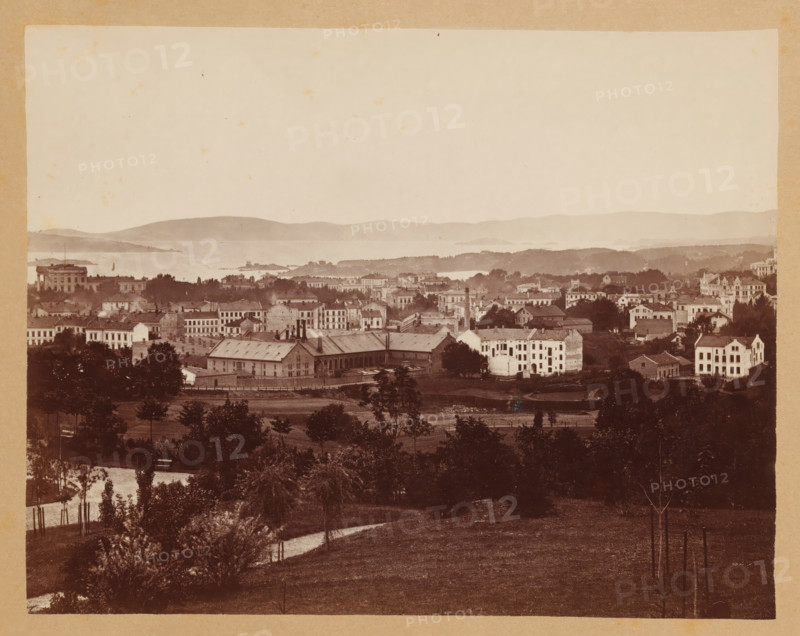 View of Christiania, Christiania, photograph, photographer: Olaf Martin Peder Væring, 1881, pasteboard, paper, albumen print, albumendruk, height 145 mm x width 183 mm, height 245 mm x width 330 mm, city-view in general, 'veduta', A sepia-toned panorama showing a townscape with rows of multi-story buildings and houses set among trees and open grassy areas, a large rectangular industrial-looking building with a sloped roof and chimneys near the center, clusters of residential structures with varied rooflines and windows spread across a gently rolling landscape, a foreground of grassy fields and scattered young trees with protective stakes and a winding dirt path, a body of water and low-lying islands visible beyond the town, and distant hazy hills forming the horizon under a pale sky.