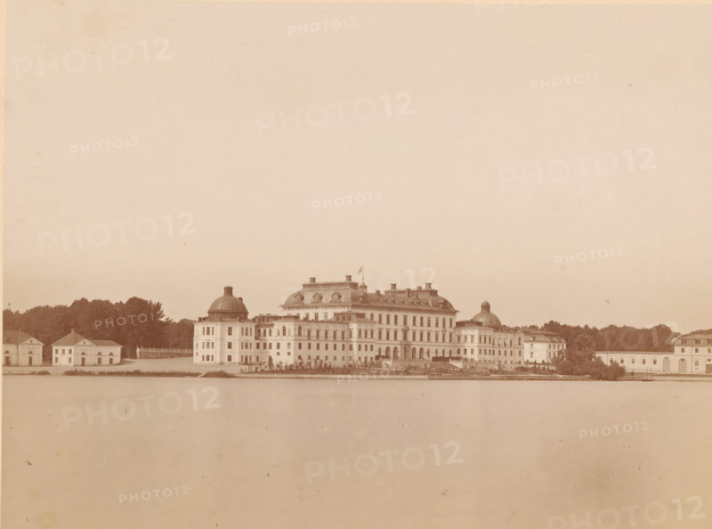 View of Drottningholm Castle near Stockholm, Drottningholms Slott, photograph, photographer: August Roesler, Stockholm, 1862 - 1893, pasteboard, paper, albumen print, albumendruk, height 161 mm x width 220 mm, height 268 mm x width 320 mm, palace, Drottningholm Castle (Ekerö), Ekerö, A wide sepia-toned photograph shows a large classical building complex along a waterfront, composed of a central grand structure with a mansard roof and ornamental rooftop elements flanked by domed pavilions and lower rectangular wings, with multiple rows of windows and arched openings on the ground level; a flag flies atop a small tower on the central roof. The complex sits behind a low waterfront embankment and a narrow strip of water in the foreground, and is set against a backdrop of dense trees and smaller outbuildings at each end. The sky above is expansive and pale, occupying most of the image, and the overall scene has an aged, uniform brownish tone with slight spots and blemishes on the surface.