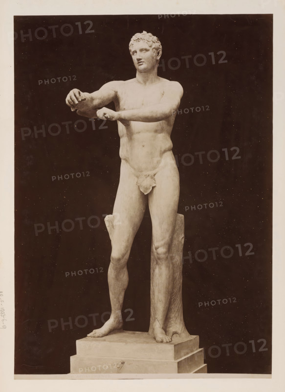 Apoxyomenos, Sculpture of Apoxyomenos, Vatican, album page, photograph, This photo is part of an album., inscription: '1315 M. Vat. APOXYOMENOS', photographer: attributed to James Anderson, Vatican Museums, after sculpture by anonymous, c. 1857 - c. 1875, after sculpture by, Vatican City, Attributed to, paper, albumen print, height 382 mm x width 273 mm, piece of sculpture, reproduction of a piece of sculpture, athletics, gymnastics, A full-length photograph of a marble male statue standing on a stepped rectangular base against a dark background, showing a nude figure with short curly hair and a calm facial expression, the torso turned slightly with both arms extended forward and hands positioned as if holding something, one forearm crossed over the other, a cloth draped over the groin, muscular legs with one foot placed forward and the other slightly back, a tree-trunk-like support behind one leg, and a smooth stone plinth beneath the feet.