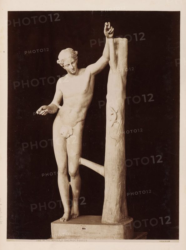 Sculpture of Apollo Sauroctonus, Vatican, Apollo Sauroctonus, album page, photograph, This photo is part of an album., inscription: '1309 M. Vat. APOLLO SAUROCTONOS Anderson', photographer: James Anderson, Vatican Museums, after sculpture by anonymous, c. 1857 - c. 1875, after sculpture by, Vatican City, paper, albumen print, height 376 mm x width 280 mm, piece of sculpture, reproduction of a piece of sculpture, A pale statue of a nude male figure stands next to a tall, textured vertical post with a horizontal support, the figure leaning with one arm raised and grasping the post while the other arm extends outward with an open hand; the figure has curly hair styled close to the head, a serene facial expression with downcast eyes, and a naturalistic, well-defined torso and limbs; the statue is mounted on a rectangular base and photographed against a dark background, with the post showing carved vine-like details and the surface of the sculpture displaying smooth and slightly weathered t...
