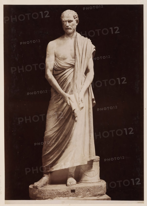 Demosthenes, Sculpture of Demosthenes, Vatican, album page, photograph, This photo is part of an album., inscription: '1358 DEMOSTENE M. Vat. Anderson', photographer: James Anderson, Vatican Museums, after sculpture by anonymous, c. 1857 - c. 1875, after sculpture by, Vatican City, paper, albumen print, height 388 mm x width 292 mm, Person, piece of sculpture, reproduction of a piece of sculpture, adult man, A full-length marble statue of a robed male figure standing on a rectangular plinth, shown against a dark background; the figure is barefoot and wears sandals, with a draped cloak over one shoulder and wrapped across the torso, and he holds a rolled object with both hands at waist level; the face has short curly hair and a beard, the expression is neutral, and the statue displays carved details of musculature, folds in the drapery, and a cylindrical support beside one leg.