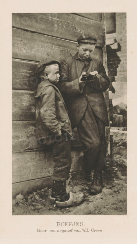 Two boys on the street, Rascals, page, photomechanical print, photographer: WL Greve, Netherlands, cliché maker: anonymous, c. 1901 - in or before 1906, paper, photoaquatint, height 139 mm x width 79 mm, boy (child between toddler and youth), child, A sepia-toned photograph of two boys standing against a weathered wooden wall, one boy leaning with his hands in his pockets wearing a cap and a jacket and looking toward the other, the taller boy standing beside him wearing a cap and a long coat while looking down at something held in his hands, both boys wearing worn trousers and boots, the shorter boy also wearing a wooden clog on one foot, rough ground with debris beneath them and a partially visible brick structure at the right edge of the image.