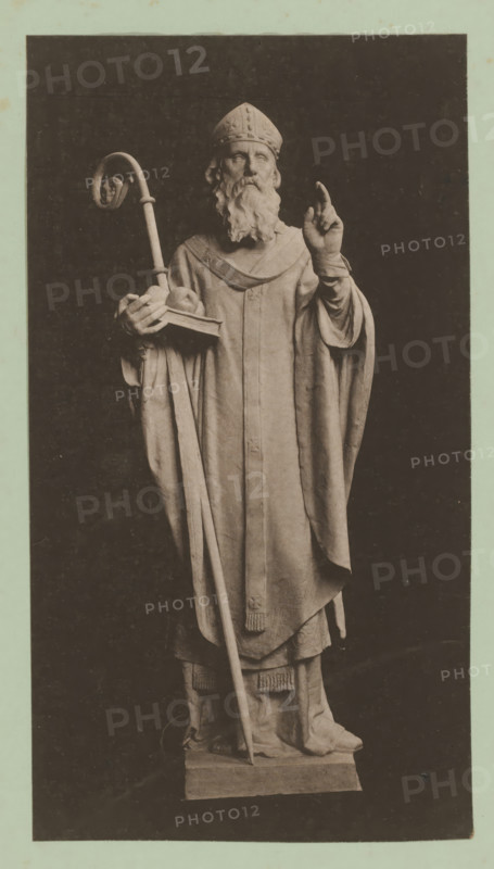 Image of a bishop, album page, photomechanical print, This print is part of an album., inscription: 'Mij. voor Photolithographr & Zincographie, procédé Mr. EJ Asser.', printer (person): Maatschappij voor Photolithographie & Zincographie, Amsterdam, 1892 - 1900, printer (person), paper, collotype, photogravure, height 281 mm x width 153 mm, archbishop, bishop, etc. (Roman Catholic), A monochrome photograph of a full-length statue of a robed figure wearing a mitre and a long flowing robe, with a long beard and mustache; the figure holds a curved staff in one hand and a book with a round object on it in the other, while the opposite hand is raised with two fingers extended; the robe features layered drapery and decorative tassels at the hem, and the statue stands on a rectangular plinth against a dark background with a pale border surrounding the image.