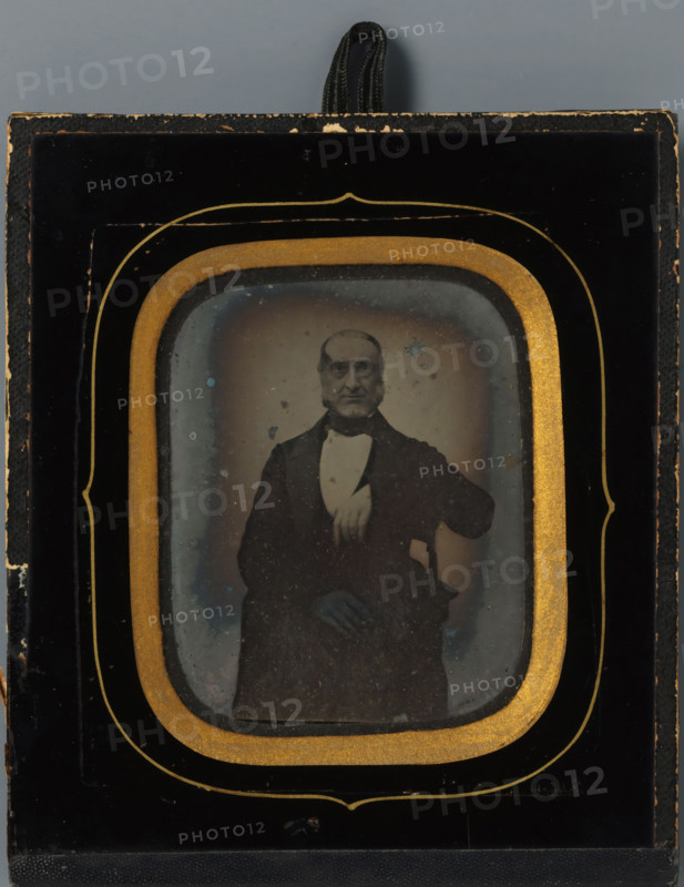 Portrait of Wouter Holleman, manufacturer, photograph, inscription: 'Wouter, 1831.', Netherlands, c. 1855 - c. 1870, photographer, pasteboard, inorganic material, glass, paper, ambrotype, height 74 mm x width 58 mm, height 129 mm x width 111 mm, Person, Wouter Holleman, historical persons, A framed photographic image shows an older man seated and facing forward, wearing a dark suit jacket with a white shirt and bow tie, one arm resting on the arm of a chair and the other hand visible near his lap; the photograph is surrounded by a decorative mat with an inner gold oval border and an outer black frame with gold trim, and a looped black cord is visible at the top of the frame.