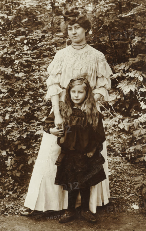 grün No 25 m. Fou ? auflage., gray at 6 fach Block, Portrait of a woman and a girl in front of a bush, photograph, annotation: 'grün No 25 m. Fou ? auflage., ... gray im 6 fach Block', photographer: attributed to Richard Tepe, Netherlands, c. 1900 - c. 1930, Attributed to, pasteboard, paper, daylight gelatin silver print, height 153 mm x width 98 mm, A sepia-toned photograph showing a woman standing behind a young girl in an outdoor leafy setting, the woman wearing a light-colored blouse with decorative smocking and voluminous sleeves and a long light skirt while the girl wears a dark dress with long sleeves and a layered skirt; the woman's hands rest gently on the girl's shoulders and the girl has long loose hair with a bow at the top, both facing forward with neutral expressions, surrounded by dense foliage and undergrowth, and both visible from head to toe with shoes on a dirt path.