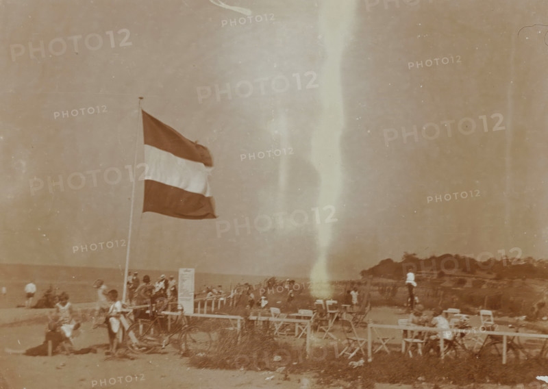 Beach at Valkeveen with the Dutch flag, photograph, inscription: 'o', Valkeveen, c. 1925 - c. 1950, photographer, paper, baryta paper, daylight collodion silver print, height 91 mm x width 119 mm, beach (as place of recreation), flag, colors (as symbol of the state, etc.), A tall flag on a pole with horizontal dark light and dark bands flutters above a sandy beach crowded with people and beach chairs, a bicycle leaning near the pole, groups of seated and standing figures wearing hats and summer clothing, rows of folding chairs and small tables in the foreground, a rocky tree-covered headland at the shoreline in the distance, and a pale vertical streak running down the center of the image.