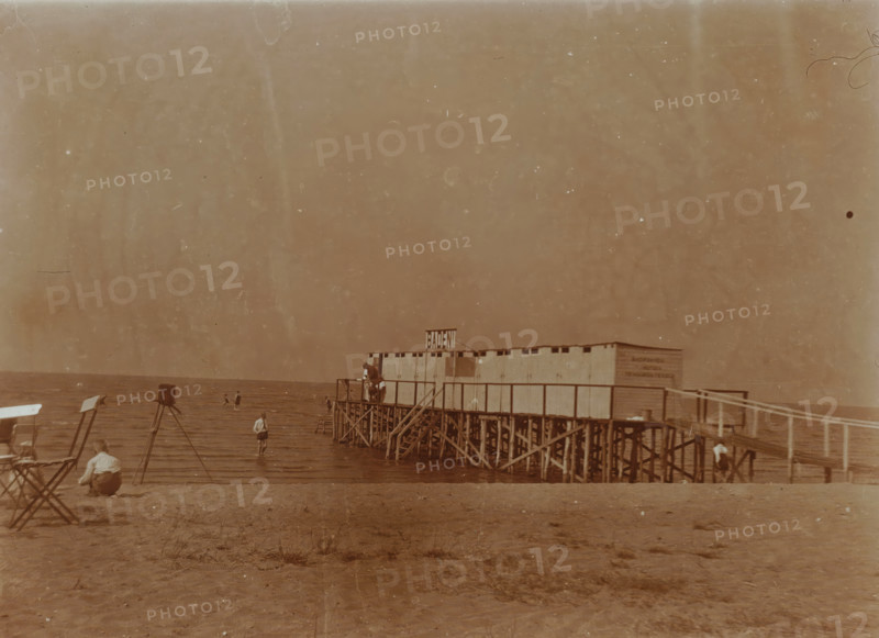 Beach at Valkeveen with changing rooms on the pier, photograph, A camera on a tripod can be seen on the beach on the left., inscription: 'Q', Valkeveen, c. 1925 - c. 1940, photographer, paper, baryta paper, daylight collodion silver print, height 91 mm x width 119 mm, beach (as place of recreation), pier, quay, wharf, A sepia-toned beach scene with a wooden pier structure extending over the water, built on a lattice of stilts and supporting a row of small changing cabins; a few people are on the pier and on the beach, some standing in shallow water and one person crouched on the sand; folding chairs and camera stands are positioned on the shore; the horizon shows calm water under a wide, empty sky.
