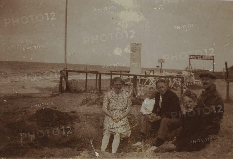 Portrait of five unknown people on the beach of Valkeveen, photograph, inscription: 'V', Valkeveen, c. 1925 - c. 1932, photographer, paper, baryta paper, daylight collodion silver print, height 59 mm x width 90 mm, beach (as place of recreation), A sepia-toned beach scene with a wooden pier extending into the sea and waves along the shoreline, worn sand and scattered seaweed in the foreground, a tall vertical post near the pier, a freestanding signboard on the beach, stacked deck chairs and a simple fence behind the group, and five people seated on the sand near the water: a woman in a patterned dress with folded hands, a small child leaning toward an older man in a dark jacket, a man wearing a cap and a coat, and another child with an open-mouthed expression.
