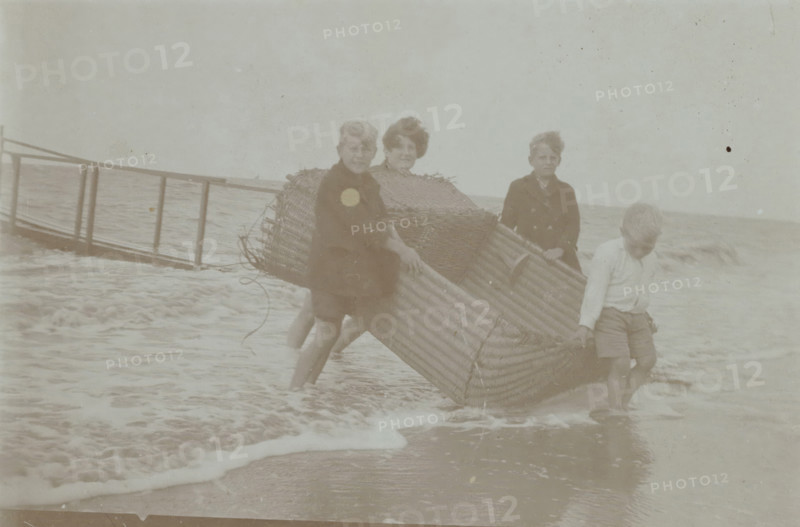 Koosje, Daan and Ben Ochse and another boy carrying a wicker beach chair out of the sea, photograph, inscription: 'AA', Valkeveen, c. 1925 - c. 1935, photographer, paper, baryta paper, daylight collodion silver print, height 59 mm x width 90 mm, Person, Koosje Ochse, Daniël Bernardus 'Ben' Ochse, Heinrich Daniël 'Daan' Ochse, beach (as place of recreation), child (four persons), A faded black and white scene at a shoreline shows four children standing in shallow water while holding a large woven wicker boat or basket. Three children face the camera, two standing close together with one partially behind the wicker, and a boy further right holding the edge and looking slightly down. Another child at the front right leans forward gripping the wicker and standing with wet legs. In the background a simple wooden railing or pier extends into the water, and small waves wash around the children and the base of the wicker, creating foamy water and wet sand at the foreground. The sky appears pal...