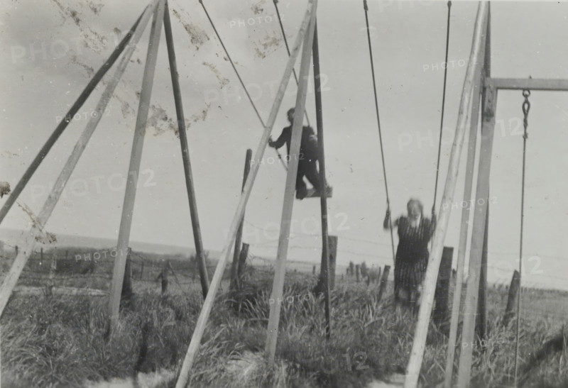 Two people swinging on the beach of Valkeveen, photograph, inscription: 'AN', number: '21', Valkeveen, c. 1925 - c. 1940, photographer, paper, baryta paper, gelatin silver print, developed gelatin silver print, height 66 mm x width 89 mm, beach, swinging games, A black and white scene showing a playground swing structure with tall wooden A-frame supports and ropes; a person sits on a raised swing seat near the center, holding ropes, and another person stands on or near a lower swing to the right, holding ropes with blurred motion; grassy ground and scattered wooden posts are visible beneath and beyond the swings, and the sky fills the upper background.