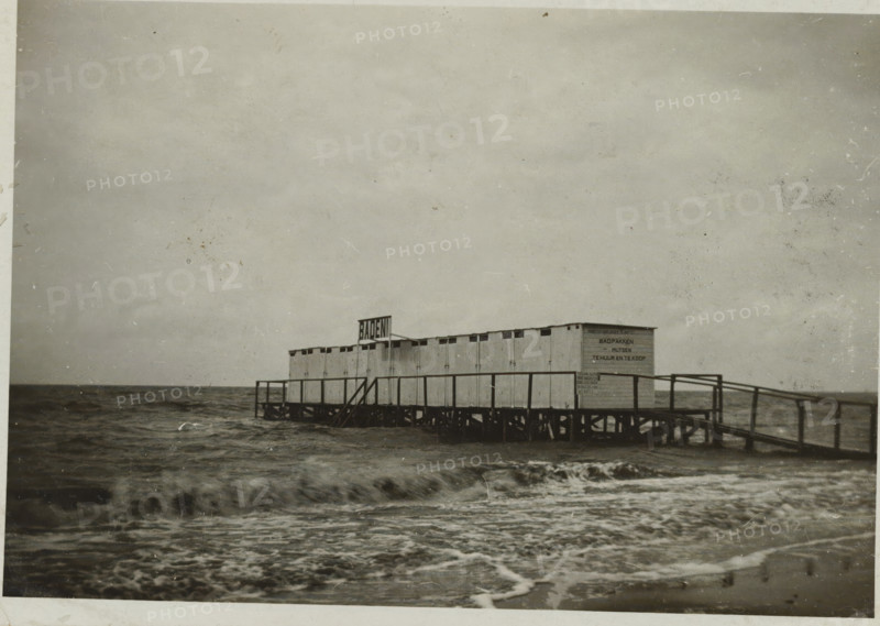 Pier with changing rooms in the Zuiderzee at the beach of Valkeveen, photograph, inscription: 'AI', Valkeveen, c. 1925 - c. 1950, photographer, paper, baryta paper, gelatin silver print, developed gelatin silver print, height 58 mm x width 82 mm, pier, quay, wharf, Zuiderzee, A long wooden pier structure with small connected cabin-like units stands on stilts over choppy dark water, a simple railing running along the pier and a set of stairs leading down toward the sea, waves breaking in the foreground and a wide expanse of cloudy sky above.
