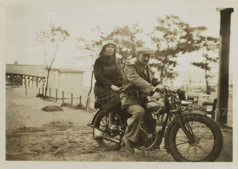 Wilhelm Ochse and Catharina Maria Groennou on a motorcycle on the beach of Valkeveen, photograph, inscription: '1934', inscription: 'AG', inscription: '4', Valkeveen, c. 1934, photographer, paper, baryta paper, gelatin silver print, developed gelatin silver print, height 60 mm x width 86 mm, Person, Catharina Maria Groennou, Wilhelm Ochse, beach (as place of recreation), motorcycles and other two-wheeled motor vehicles, A sepia-toned photo shows two people seated on an old motorcycle on a sandy surface near a wooden building and a raised pier, with trees and sparse vegetation in the background; the person in front wears a flat cap and a coat and has one hand on the handlebar while the person behind wears a cloche-style hat and a long coat and holds onto the seat, the motorcycle's visible parts include a front wheel with spokes, a headlamp, an engine, and a license plate mounted at the front, and a vertical wooden post appears at the right edge of the image.