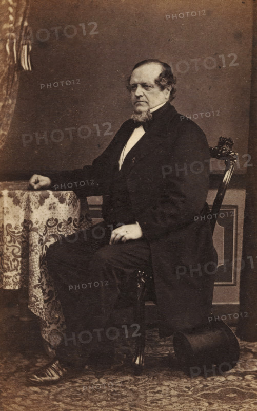 Portrait of an unknown man from the Marmelstein family, photograph, carte-de-visite, annotation: 'From Fr. F', annotation: 'Grandfather Blok, Scato Trip', 1850 - 1900, photographer, paper, pasteboard, albumen print, height 93 mm x width 60 mm, historical persons not known by name, A seated older man wearing a dark formal coat and light shirt with a bow at the collar sits on an ornately carved wooden chair, one arm resting on a table covered with a patterned cloth and the other hand resting on his knee; he has a full beard along his chin and short hair, and a top hat rests on the floor beside the chair on a patterned rug, with a plain backdrop behind them.