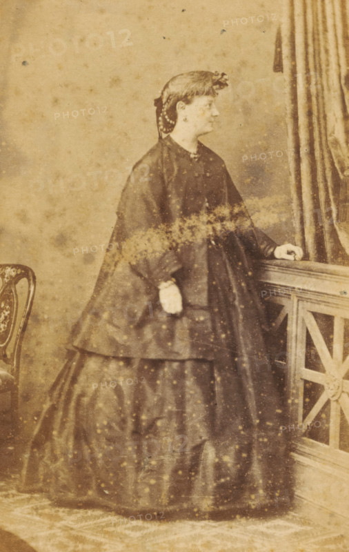 Portrait of an unknown woman from the Marmelstein family, photograph, carte-de-visite, photographer: Tresling & Comp., Amsterdam, 1850 - 1900, paper, pasteboard, albumen print, albumendruk, height 106 mm x width 61 mm, historical persons, adult woman, A sepia-toned portrait of a woman standing in profile facing right, wearing a dark full-skirted dress with layered fabric and a fitted bodice, a cape-like outer garment, and light-colored gloves; her hair is styled and adorned with a ribbon or decorative piece, and she has earrings and a necklace; she rests one hand on a wooden railing with a geometric carved panel while the other hand hangs by her side; a patterned chair is partially visible at the left edge and a heavy drapery hangs at the right, the background is plain with visible age spots and marks across the image.