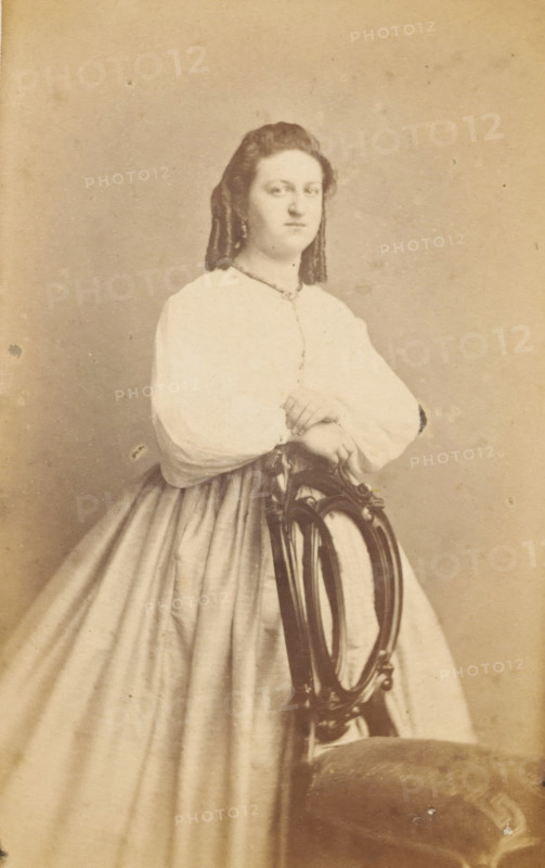 Portrait of an unknown woman from the Marmelstein family, photograph, carte-de-visite, inscription: 'J. KORSTEN, Painter, and PHOTOGRAPH, MIDDELBURG.', photographer: J. Korsten, Middelburg, 1864 - 1878, paper, pasteboard, albumen print, albumendruk, height 103 mm x width 61 mm, historical persons - BB - woman, A monochrome portrait of a woman standing and leaning with her folded hands on the back of an ornate wooden chair, wearing a light-colored dress with a full skirt and long sleeves, her hair styled away from her face and falling in waves, a neutral facial expression, and a plain backdrop behind her.