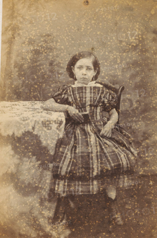 Portrait of an unknown girl from the Marmelstein family, photograph, carte-de-visite, address: 'PHOTOGRAPHISCH ATELIER., J. WIJNMAN, Halsteeg No. 248., Amsterdam.', photographer: J. Wijnman, Amsterdam, 1866 - 1870, paper, pasteboard, albumen print, albumendruk, height 108 mm x width 64 mm, historical persons not known by name - BB - woman, child, A seated young person with a solemn facial expression wears a striped dress with puffed sleeves and a white collar, one arm resting on a lace-covered table and the other hand holding the skirt; dark hair is styled close to the head and the figure sits on a wooden chair with a curved back, against a mottled backdrop with numerous light speckles and age spots visible across the image.