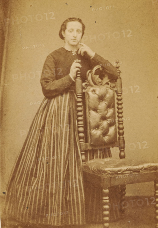 Portrait of an unknown woman from the Marmelstein family, photograph, carte-de-visite, photographer: Caspel & Stapert, S. van, Amsterdam, 1850 - 1900, paper, pasteboard, albumen print, albumendruk, height 110 mm x width 65 mm, historical persons, adult woman, A sepia-toned photograph shows a person standing beside an ornate wooden chair with a tufted upholstered back and seat; the person wears a dark long-sleeved bodice with a light collar and a long skirt with vertical stripes, has hair pulled back, and rests one hand on the top of the chair while the other hand is raised near the face.