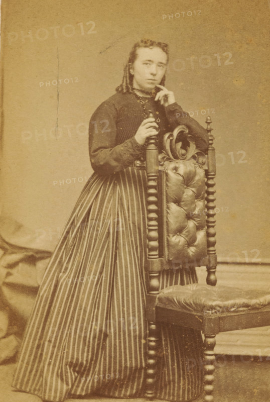 Portrait of an unknown woman from the Marmelstein family, photograph, carte-de-visite, photographer: Caspel & Stapert, S. van, Amsterdam, 1850 - 1900, paper, pasteboard, albumen print, albumendruk, height 111 mm x width 65 mm, historical persons, adult woman, A sepia-toned portrait of a person standing beside an ornate wooden chair with a tufted leather back and seat, the person wearing a dark long-sleeved bodice and a long skirt with vertical stripes; one hand rests on the turned finial of the chair while the other hand is raised near the face, and the person looks slightly forward with a neutral expression, hair pulled back and styled; the background is plain and textured.