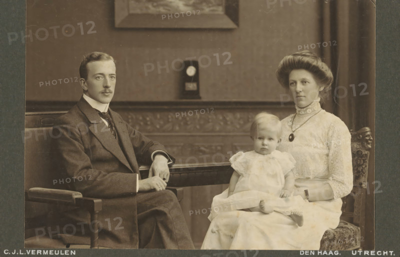 Portrait of an unknown couple and their daughter, cabinet photograph, inscription: 'CJL Vermeulen, Studio for modern photography, The Hague, Toussaintkade 11, Telephone 1873., Utrecht, Nieuwegracht 50, Telephone 750.', annotation: 'Rijswijk 1911.', photographer: Cornelis Johannes Lodewicus Vermeulen, Netherlands, 1890 - 1930, paper, pasteboard, daylight collodion silver print, height 108 mm x width 165 mm, historical persons not known by name, historical persons not known by name - BB - woman, child, A sepia-toned portrait of a family seated indoors on carved wooden chairs, showing a man in a dark suit with a high collar and tie sitting on the left, a woman in a light lace dress with a high lace collar and a pendant necklace seated on the right, and a small child in a light frilly dress sitting on the woman’s lap; the man has a neatly trimmed mustache and rests one arm on the chair arm while the other hand is near his knee, the woman has an upswept hairstyle and a composed expression, ...