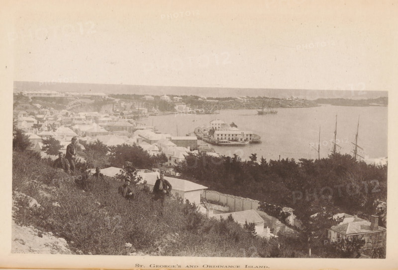 St. George's Island and Ordinance Island, page, photomechanical print, photographer: James H. Stark, Bermuda, cliché maker: Photo-Electrotype Company, Boston (Massachusetts), c. 1879 - in or before 1884, James H. Stark, Photo-Electrotype Company, photographer, cliché maker, North America, Massachusetts, paper, collotype, photoprint, height 99 mm x width 152 mm, island, Saint George's Island, Ordnance Island, A sepia-toned coastal scene showing a hillside covered in scrub and bushes descending to a settlement of light-roofed buildings and houses clustered along a shoreline and extending inland, with a harbor area containing piers and a wharf where a multi-deck vessel is moored, and tall masts of sailing ships visible near the water; several figures in period clothing stand or sit on the hillside among the vegetation, and trees and larger shrubs frame parts of the foreground and midground while a distant low shoreline and calm water stretch across the background under a pale sky.