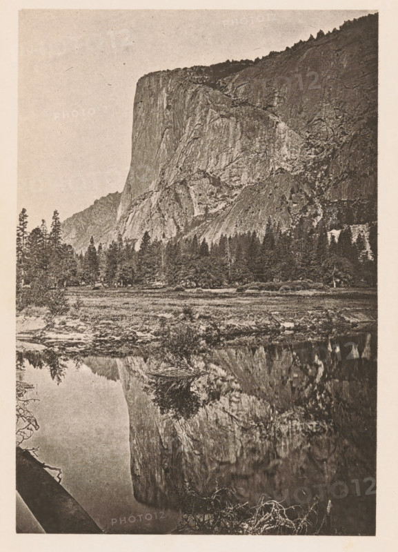 El Capitan, Yosemite Valley. 3, 300 feet high, View of El Capitan in the Yosemite Valley, page, photomechanical print, Yosemite Valley, c. 1866 - in or before 1876, North America, California, paper, photoaquatint, height 157 mm x width 108 mm, Print opposite title page., rock-formations, A vertical monochrome photograph showing a steep rocky cliff rising from a forested foothill, with rugged rock faces and textured slopes; a line of pine trees stands at the base of the cliff, and a grassy meadow lies between the trees and a calm reflective body of water in the foreground that mirrors the cliff and some trees; the sky above is clear and pale, and the image has a grainy, aged paper appearance with a narrow light border around it.
