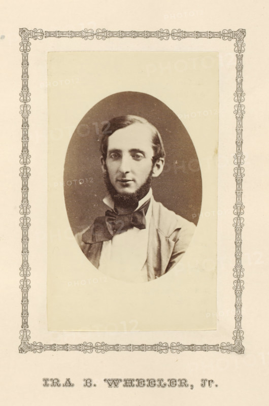 Ira B. Wheeler, Jr., photograph, page, c. 1876 - in or before 1881, photographer, paper, albumen print, height 100 mm x width 60 mm, adult man, A sepia-toned oval portrait of a young man with short dark hair and sideburns, wearing a light-colored shirt, a dark bow tied at his collar, and a jacket, set on a pale rectangular mount with a decorative border surrounding the mount and the page.