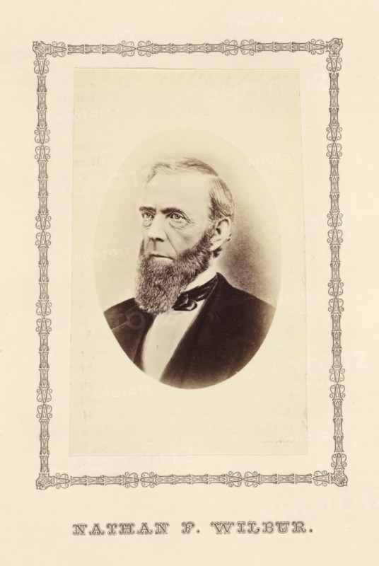 Nathan F. Wilbur, photograph, page, c. 1876 - in or before 1881, photographer, paper, albumen print, height 100 mm x width 60 mm, adult man, A sepia-toned oval portrait of a man with a full beard and neatly combed hair, shown from the chest up and dressed in a dark coat with a light shirt and a bow at the collar, centered on a rectangular off-white page with an ornate decorative border surrounding the portrait.