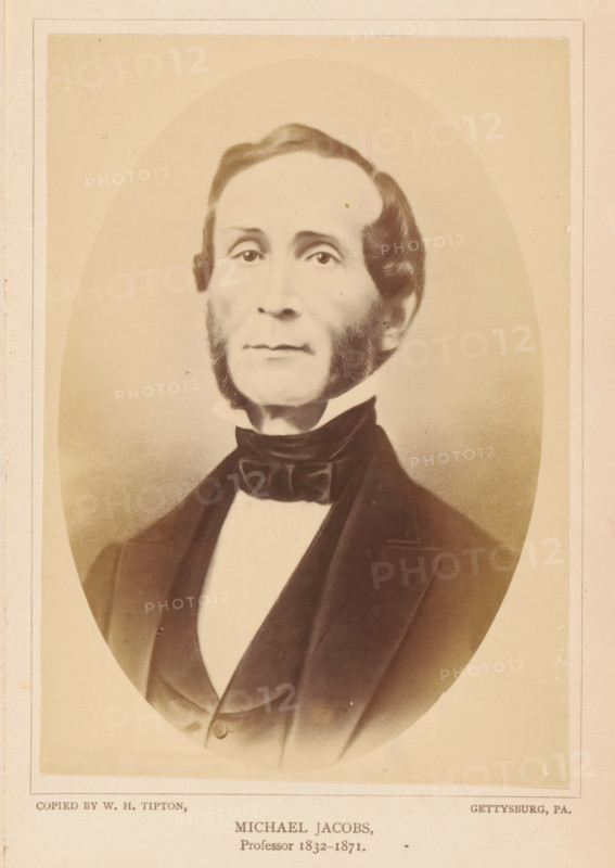Michael Jacobs, photograph, reproduction, page, photographer: William H. Tipton, Gettysburg, after drawing by anonymous, c. 1877 - in or before 1882, North America, Pennsylvania, paper, albumen print, height 142 mm x width 103 mm, Person, professor, A sepia-toned oval portrait of a middle-aged man shown from the chest up, centered on a pale background with a rectangular border. He has a high forehead with neatly combed hair that parts to one side and rounded sideburns framing his cheeks and jaw. His expression is neutral, with a straight mouth and eyes looking forward. He wears a dark formal jacket over a light shirt with a high collar and a dark bow tied around the neck. The image shows subtle shading and photographic texture, with the edges of the oval fading into the surrounding background.