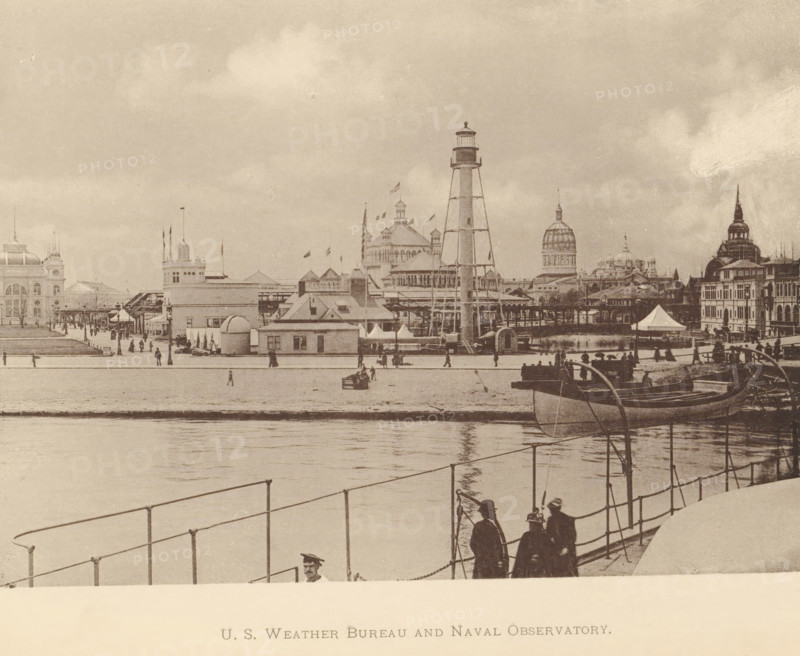 Coast Guard station and weather bureau at the World's Columbian Exposition in Chicago in 1893, US Weather Bureau and Naval Observatory, page, photomechanical print, Chicago, 1893, North America, Illinois, paper, collotype, photoprint, height 128 mm x width 177 mm, rowing-boat, canoe, world fair, world exhibition, World's Columbian Exposition, A waterfront scene in a sepia photograph shows a pier area with a railing in the foreground and calm water reflecting the shore. Several people stand near the railing, some wearing hats and coats, and a small group cluster together. Boats are moored along the shore, one prominent boat angled toward the viewer. On the opposite bank a row of buildings and pavilions rise, including a tall skeletal lighthouse tower, a domed building, and structures with turrets and flagpoles. Tents and smaller booths line the shore and a promenade runs along the water with scattered pedestrians. The sky above is cloudy and the overall image has an aged, grainy texture...