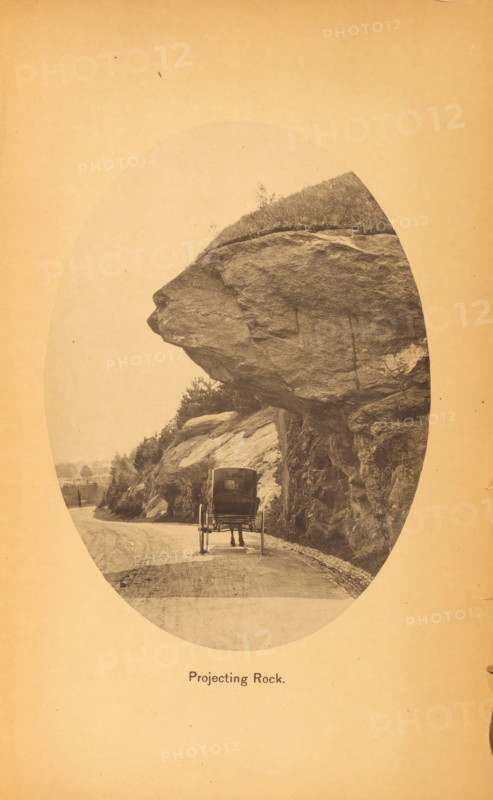 Projecting rock, page, photomechanical print, New York (city), c. 1866 - in or before 1871, North America, paper, collotype, photoprint, height 160 mm x width 116 mm, Print after page 157., public road, four-wheeled, animal-drawn vehicle, eg: cab, carriage, coach, rock-formations, A sepia-toned oval photograph shows a dirt road curving beside a large rock face with a massive overhanging ledge protruding above the roadway; beneath the ledge a horse-drawn carriage faces away from the viewer with the horse partially visible and the carriage wheels and rear seen; the rock surface displays rough textures and strata with vegetation and small shrubs on top and along the slope; the road edge is lined with small stones and the distant background shows faint trees and landscape.