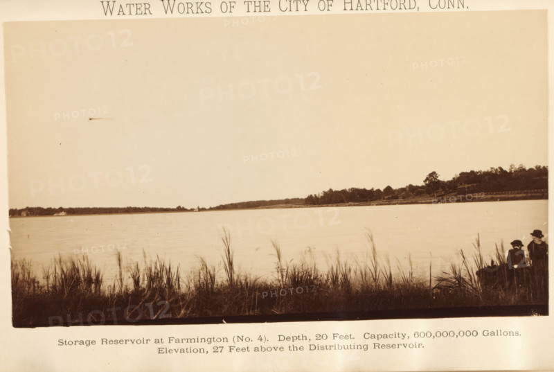 Water works of the city of Hartford, Conn, Storage reservoir at West Hartford (no. 4). Depth, 20 feet. Capacity 600, 000, 000 gallons. Elevation, 27 feet above Connecticut river, photograph, page, Connecticut, c. 1883 - in or before 1884, photographer, North America, paper, albumen print, height 108 mm x width 180 mm, water course, A wide sepia-toned photograph showing a calm body of water extending toward a distant tree-lined shoreline, with tall grasses and reeds in the foreground along the near bank; on the right edge two seated figures wearing dark clothing and hats are partially visible among the grasses; the sky above the water is mostly clear and light in tone.