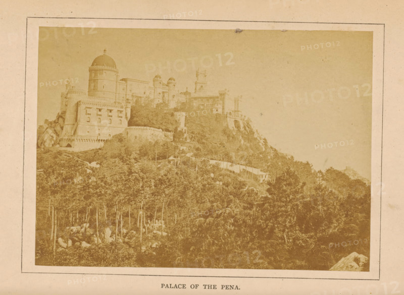 View of the Palácio da Pena in Sintra, Palace of the Pena, photograph, page, Sintra, c. 1860 - in or before 1870, photographer, paper, albumen print, height 97 mm x width 135 mm, palace, Palácio da Pena, A faded photograph mounted on a pale mat shows a hillside topped by a large ornate building with rounded towers, domes, battlements, arched openings, and decorative stonework, with a dense stand of trees and rocky terrain covering the slope below and a lighter sky occupying the upper right area.