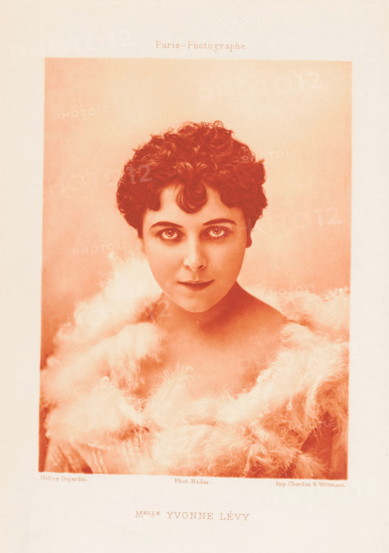 Portrait of Yvonne Lévy, Melle Yvonne Lévy, page, photomechanical print, photographer: Félix Nadar, Paris, cliché maker: Dujardin, printer (person): Chardon-Wittmann, c. 1882 - in or before 1892, printer (person), paper, photoaquatint, height 143 mm x width 106 mm, Person, Yvonne Lévy, portrait of actor, actress, A portrait of a young woman with short curly hair framed in a soft rectangular vignette, shown from the shoulders up and wearing a fluffy, feathery wrap around her shoulders and neck, with large expressive eyes looking toward the viewer and a neutral mouth, rendered in a warm monochrome tone against a plain background.