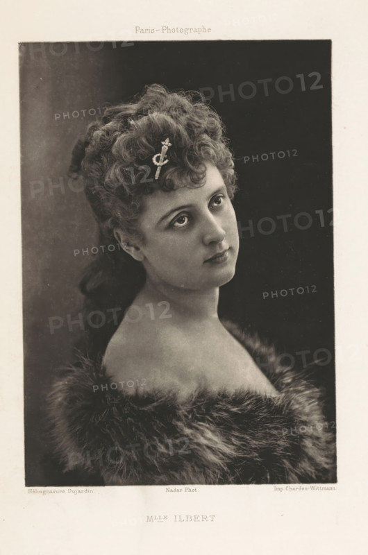 Mlle Ilbert, Portrait of Yvette Guilbert, page, photomechanical print, photographer: Félix Nadar, Paris, cliché maker: Dujardin, France, printer (person): Chardon-Wittmann, c. 1882 - in or before 1892, printer (person), paper, photoaquatint, height 159 mm x width 112 mm, Person, Yvette Guilbert, female singer, A sepia-toned portrait of a young woman shown from the shoulders up, her head tilted slightly and gaze directed upward and to the side, with tightly curled hair gathered and styled at the top and back, a decorative hairpin placed among the curls, clear eyes and delicate facial features, bare shoulders framed by a soft, plush fur wrap, and a plain dark backdrop inside a bordered rectangular frame on a light paper mount.