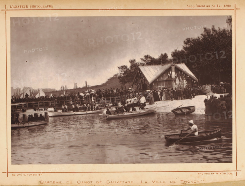 Baptism of the lifeboat La ville de Thonon, Baptême du canot de sauvetage 'La ville de Thonon', page, photomechanical print, photographer: E. Forestier, cliché maker: Adolphe Block, c. 1885 - in or before 1890, paper, Woodburytype, height 111 mm x width 168 mm, lifeboat, christening a ship, A sepia-toned scene shows a waterfront with several small rowboats on calm water, one in the foreground with a person wearing a hat rowing, and other boats nearer the shore with people seated or preparing to row. A wooden pier crowded with people extends along the shoreline, and a group of people stands on the sandy beach area near a pavilion-style wooden building decorated with hanging flags and trim. Trees rise behind the building, and additional crowds gather near the treeline and along the pier. Sails and masts are visible in the distance behind the pier, and the sky above is overcast.