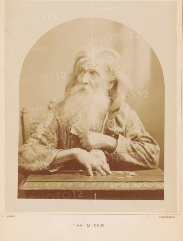 The Miser, Portrait of an Unknown Old Man, photograph, page, photographer: James M. Landy, Cincinnati (possibly), printer (person): Fellows & Graves, Philadelphia, c. 1887 - in or before 1892-12-03, printer (person), North America, paper, albumen print, height 161 mm x width 131 mm, historical persons not known by name, old man, An old man with long white hair and a long white beard sits behind a carved table, wearing a patterned robe and gripping a cloth near his chest while his other hand rests on the table with fingers splayed over several round objects; he looks upward and slightly to the side, and the photograph shows an arched vignette around him with a visible vertical crease running through the center of the image.
