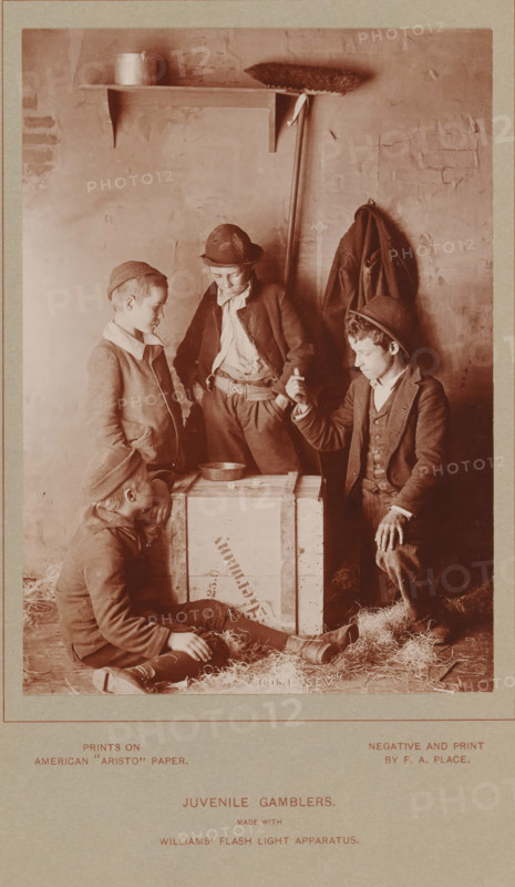 Juvenile Gamblers, Four unknown boys playing dice, photograph, page, inscription: 'made with, Williams' flash light apparatus.', inscription: 'Prints on, American 'Aristo' Paper', photographer: Francis A. Place, c. 1888 - in or before 1893, Francis A. Place, photographer, paper, daylight collodion silver print, height 140 mm x width 99 mm, Photo two pages before page 142., child, dice, A sepia photograph shows four boys gathered around a wooden crate in a dim interior space with a rough plastered wall. One boy sits on the floor leaning against the crate with his legs extended and a cap on his head, another stands to the left with hands in his pockets looking toward the center, a third stands behind the crate with a hand in his pocket wearing a hat and looking down, and a fourth kneels to the right holding a small object between his fingers while looking at it. A shallow metal bowl rests on top of the crate, straw is scattered on the floor, a coat hangs on the wall behind them, and a br...