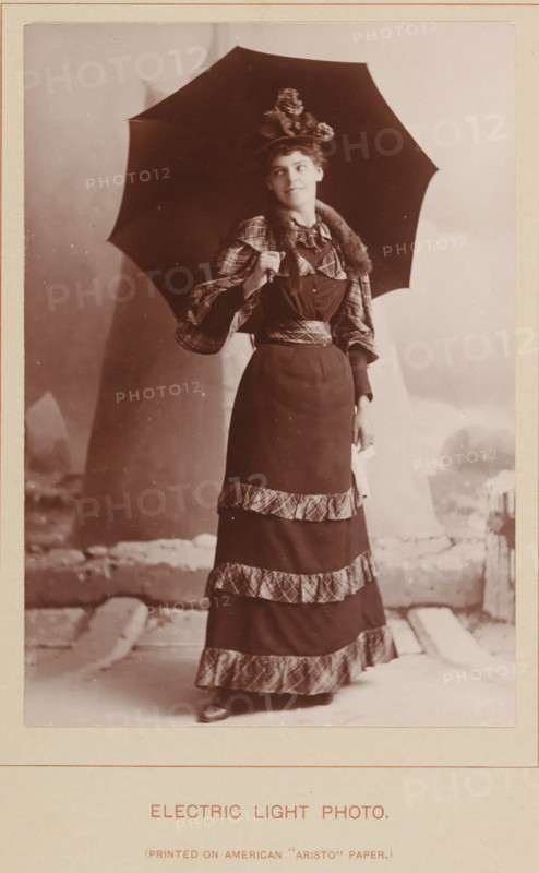 Electric Light Photo, Portrait of an unknown woman with an umbrella, photograph, page, inscription: 'Pinted on American 'Aristo' Paper', c. 1888 - in or before 1893, photographer, paper, daylight collodion silver print, height 140 mm x width 98 mm, Photo two pages before page 178., adult woman, A sepia-toned portrait of a woman standing and holding a large dark umbrella over her shoulder, wearing a long dark dress with patterned trim bands on the skirt and matching patterned sleeves, a fur or furlike collar, and a decorative hat with flowers, her hair styled up and her face turned slightly to one side with a faint smile, one hand grasping the umbrella handle and the other holding a light-colored cloth, with a studio backdrop and a visible floor area beneath her feet.