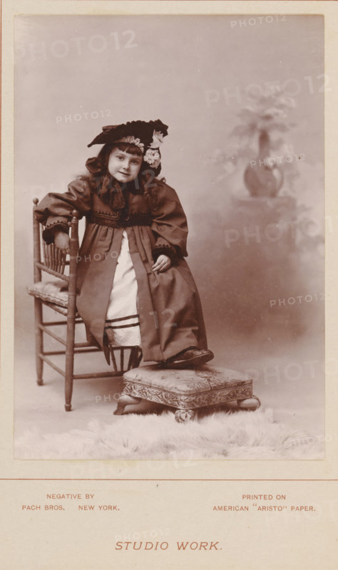 Portrait of an unknown girl on a chair, Studio work, photograph, page, inscription: 'printed on American 'Aristo' paper', photographer: Pach Brothers, c. 1888 - in or before 1893, paper, daylight collodion silver print, height 140 mm x width 99 mm, Photo two pages before page 246., historical persons, girl (child between toddler and youth), chair, A sepia photograph of a young girl seated on a wooden chair, wearing a long dress with a lighter apron-like panel and a wide-sleeved coat, a large decorative hat with floral adornments, and shoes; one foot rests on a patterned cushioned footstool placed on a furry rug, and a blurred vase with flowers appears in the background.