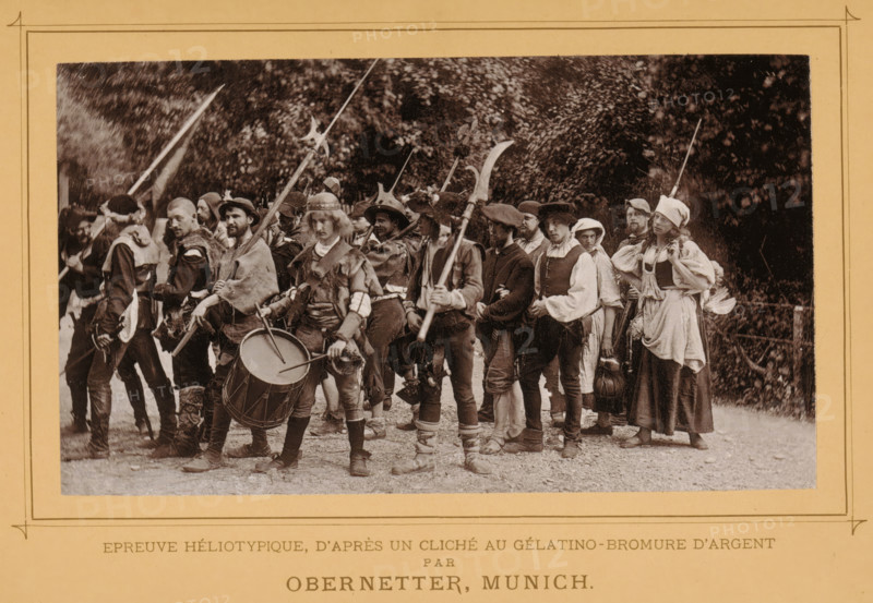 View of a group of unknown persons, one with a drum, probably re-enacting a fight or battle, page, photomechanical print, inscription: 'Épreuve heliotypique, d'après un cliché au gélatino-bromure d'argent, par, Obernetter, Munich', cliché maker: Johann Baptist Obernetter, c. 1884 - in or before 1889, photographer, printer (person), paper, collotype, photoprint, height 96 mm x width 166 mm, Print two pages after page 318., historical persons, drum (musical instrument) - CC - out of doors, A sepia-toned photograph shows a group of people standing outdoors in a line, many wearing historical-style clothing and hats, with some holding long pole weapons and one person holding two drumsticks over a drum; men and women are visible, dressed in layered garments with cloaks, tunics, and head coverings, and several have boots and wrapped leg coverings; faces show neutral or focused expressions and hairstyles range from short to shoulder-length; trees and foliage form a dark backdrop behind the gro...