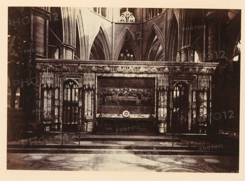 Altarpiece at Westminster Abbey, photograph, page, photographer: John Harrington, c. 1864 - in or before 1869, paper, albumen print, height 130 mm x width 186 mm, interior of church, abbey, monastery, convent, Roman Catholic church, altar with dossel (dorsal), 'reredos', altar-screen, retable, London, Westminster Abbey, A wide interior view of an ornate stone structure with tall pointed arches and ribbed vaulted ceilings behind a carved marble screen, featuring decorative columns and sculpted relief along its upper edge. In the central opening of the screen there is a framed panel depicting a seated figure surrounded by others at a long table, set above an intricately carved base. Flanking the central panel are two tall arched openings with tracery and clustered slender columns, and above the screen a row of sculpted roundels and ornamental figures runs horizontally. The foreground shows a patterned stone floor with circular motifs and a low railing with metal posts, while the sides of...