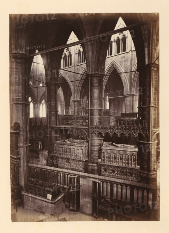 Chapel of St. Edmund at Westminster Abbey, photograph, page, photographer: John Harrington, c. 1864 - in or before 1869, paper, albumen print, height 194 mm x width 139 mm, interior of church, abbey, monastery, convent, Roman Catholic church, parts of church interior: chapel, London, Westminster Abbey, The image shows the interior of a large stone building with tall, ribbed pointed arches and clustered columns supporting vaulted ceilings; ornate carved stone screens and tombs with recumbent effigies sit within the space, and a decorative railing and intricate sculpted panels are visible in the foreground; light streams through tall arched windows in the background, casting soft highlights on the stone surfaces and emphasizing the vertical architectural elements.