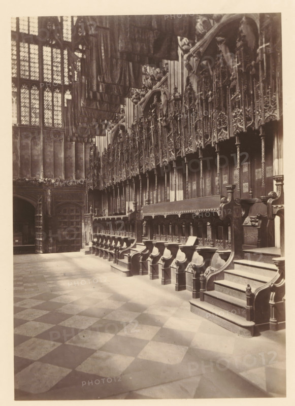 Chapel of Henry VII at Westminster Abbey, photograph, page, photographer: John Harrington, c. 1864 - in or before 1869, paper, albumen print, height 177 mm x width 125 mm, interior of church, abbey, monastery, convent, Roman Catholic church, parts of church interior: chapel, London, Westminster Abbey, A sepia-toned interior photograph showing a long row of ornately carved wooden choir stalls with high backs and armrests, a stepped platform at one end, and decorative panels and vertical columns above them; a patterned stone floor of alternating light and dark squares stretches toward the foreground; tall leaded glass windows with geometric panes occupy the left wall, and heavy hanging textiles or banners are visible near the ceiling; stone walls and an archway with a large wooden door appear at the far left, and detailed woodwork and tracery fill the upper right area of the image.