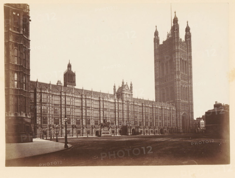 View of Palace of Westminster, south view, photograph, page, photographer: John Harrington, London, c. 1864 - in or before 1869, paper, albumen print, height 141 mm x width 195 mm, palace, Houses of Parliament, A wide sepia photograph of an ornate stone building with a long facade featuring many tall, narrow windows and decorative vertical and horizontal detailing, ending at a very tall, square tower with pinnacles and arched openings, several smaller towers and turrets rising along the roofline, a large arched entrance at the base of the tall tower, a clear paved area and roadway in front, period-style lampposts lining the pavement, a mounted statue on a plinth near the center of the facade, and a smaller cluster of buildings visible at the far right edge.