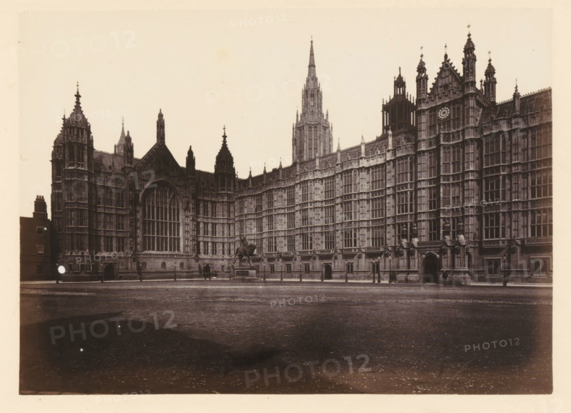 View of Palace of Westminster, north view, photograph, page, photographer: John Harrington, London, c. 1864 - in or before 1869, paper, albumen print, height 139 mm x width 195 mm, palace, Houses of Parliament, A wide sepia photograph shows an ornate Gothic stone building with tall pointed spires, decorative pinnacles, and many vertical windows set in a long facade; a large arched window with intricate tracery is prominent near the left, and a clock face is visible on a tower toward the right; a statue of a mounted figure stands on a pedestal in front of the building, with a paved open area stretching across the foreground and a few lampposts and small doorways along the base of the structure.