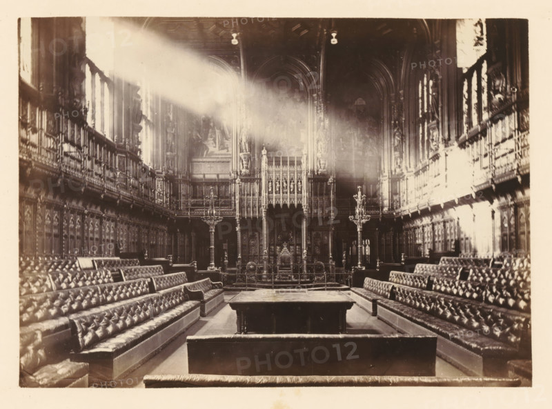 View of the interior of the Commons Chamber, photograph, page, photographer: John Harrington, London, c. 1864 - in or before 1869, paper, albumen print, height 139 mm x width 195 mm, palace, interior, representation of a building, Houses of Parliament, A wide interior view of a grand hall with high vaulted ceilings and tall windows along both side walls, sunlight streaming in as a bright diagonal beam. The room contains long, upholstered benches arranged in parallel rows facing a raised ornate screen at the far end, which features intricate carved panels, statues, and decorative columns. Two tall, ornate candle stands flank the central aisle near the screen. The side walls are lined with elaborate wood paneling and carved figures set into niches above the panels. The ceiling shows decorative moulding and hanging light fixtures. The overall tone of the image is sepia, and the floor appears smooth and reflective in places.