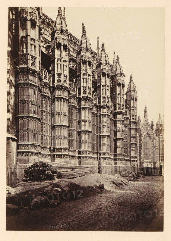 View of Westminster Abbey, Chapel of Henri VII, photograph, page, photographer: John Harrington, c. 1864 - in or before 1869, paper, albumen print, height 177 mm x width 125 mm, church (exterior), abbey, monastery, convent, Roman Catholic church, London, Westminster Abbey, A sepia-toned photograph of a large ornate stone building with tall vertical buttresses and intricately carved Gothic detailing along its façade, showing rows of deep-set windows with tracery and decorative pinnacles rising from the roofline; a cobblestone street runs alongside the building with a pile of long wooden beams and stacked logs near a low fence, and a mound of earth and scattered stones in the foreground; further along the building a large pointed arched window and additional spires are visible, and a narrow strip of pale sky fills the upper right portion of the image.